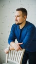 A young man leaning on the back of a chair looks to the side against a white brick wall