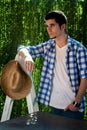 Young man leaning on a garden ladder wearing a straw hat, with a green background of plants behind