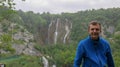 A young man leans on a wooden railing. In the background waterfalls in Plitvice Lakes National Park. Teenager in a blue hoodie