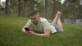 A young man lies on the grass and texts in a smartphone. Satisfied man is resting in nature with a phone. Royalty Free Stock Photo