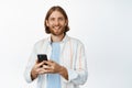 Young man listening music on headphones, smiling, holding mobile phone, using earphones, standing against white Royalty Free Stock Photo