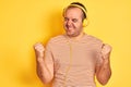 Young man listening to music using headphones standing over isolated yellow background very happy and excited doing winner gesture Royalty Free Stock Photo