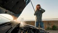 Young man looking desperate, using smartphone, calling for assistance while standing near his broken car with open hood Royalty Free Stock Photo