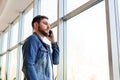 Young man making business call talking by phone indoors near window wall in casual jean jacket. Smart man in modern city office Royalty Free Stock Photo