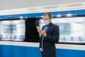 Young man manager uses cellphone, prevents spreading of Coronavirus, poses against subway train, poses at platform, checks news Royalty Free Stock Photo