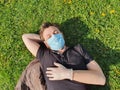 Caucasian young man with medical face mask and medical gloves lies in the grass in the park. Royalty Free Stock Photo