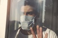 Young man in medical mask is looking out window. Coronavirus pandemic. Home quarantine, self-isolation because of the Coronavirus