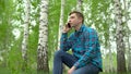 Young man in nature with a phone. A man sits on a stump in a birch forest and talks on the phone.