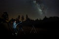 Young man observes starry sky through a telescope. Mountains, surrouded by pine tree forest in the background night landscape with Royalty Free Stock Photo