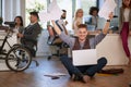 Young man at the office sitting o n the floor with his laptop with hands holding papers in the air after receiving good business