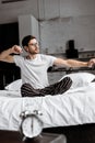 Young man in pajamas and eyeglasses stretching while sitting on bed Royalty Free Stock Photo