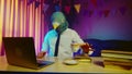 A young man in a pigeon mask is sitting on a chair at a table and typing on a laptop keyboard. A man in a mask drinks Royalty Free Stock Photo