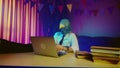 A young man in a pigeon mask is sitting on a chair at a table typing on a laptop keyboard. Multicolored lighting Royalty Free Stock Photo