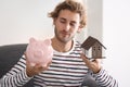 Young man with piggy bank and house model sitting in armchair at home Royalty Free Stock Photo