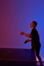 Young man, player ready to hit ball in neon-lit table tennis match against gradient pink-blue background. Dynamic gel