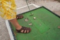 Young man playing minigolf on old, worn hotel resort green field, closeup to his feet in sandals, metal club and ball Royalty Free Stock Photo