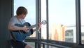 Young man plays guitar at home. Concept. Student plays black guitar standing on balcony. Guitarist plays through window Royalty Free Stock Photo