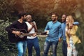 A young man plays the guitar in a multiethnic company of five people. Friends having fun in a park during sunset. Impromptu jam