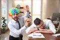 Young man popping paper bag behind his sleeping colleague in office. Funny joke