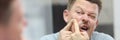 Young man presses pimple while standing in front of mirror in bathroom