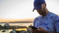 young man with a purple cap watching the smartphone at a beautiful view of the sea at the sunset Royalty Free Stock Photo