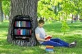 Young man reading a book sitting on the green grass leaning on a tree in the Park.