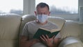 Young man reading a book sitting in a medical mask at home on the couch