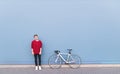 Young man in a red sweatshirt standing beside a white bicycle on a blue background and looking into the camera Royalty Free Stock Photo