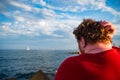 Young Man In Red T-shirt With Psoriasis Sits At Sunset On The Pier Of The Balearic Sea, Scratches His Head Thoughtfully And Looks