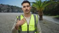 Young man with reflective vest stands outdoors in a park, displaying a thoughtful expression against a backdrop of palm trees and Royalty Free Stock Photo