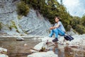 Young man relaxing by mountain river enjoying natural landscape. Traveller backpacker sitting on rock. Summer trip