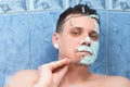 Young man is removing alginate mask from his face lying in bathroom with foam.