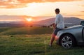 Young man resting during long solo auto trip sitting on his luxury car bonnet and enjoying the sunset sky colors. Traveling by car Royalty Free Stock Photo