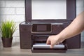 A young man`s hand places a stack of white paper in the print tray of a black printer. Green plant in a pot on a wooden Royalty Free Stock Photo