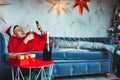 Young man in Santa Claus hat browses smartphone and drinks champagne, lying on couch. Handsome guy resting with mobile phone and Royalty Free Stock Photo