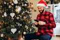 Young man in Santa Claus hat, uses a mobile phone, dials sms, has merry Christmas, sitting near a Christmas tree at home Royalty Free Stock Photo
