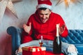 Young man in Santa hat celebrating Christmas in solitude at home. Adult guy rests, sitting on sofa in room. Concept of Royalty Free Stock Photo