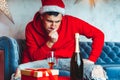 Young man in Santa hat celebrating Christmas in solitude at home. Adult guy rests, sitting on sofa in room. Concept of Royalty Free Stock Photo