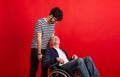 Young man and senior father in wheelchair in a studio on red background. Royalty Free Stock Photo