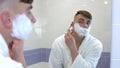 A young man shaves his face hair in front of a mirror. A man in a white coat with foam on his face shaves his hair. View Royalty Free Stock Photo