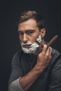 Young man with shaving cream on his face, grooming his beard with straight razor and looking Royalty Free Stock Photo