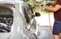 Young man in shorts and t shirt washing his car in self serve carwash, cleaning side windows with brush