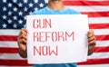 young man showing Gun reform Now sign board in front of US american flag - concept of protest against gun violence and Royalty Free Stock Photo