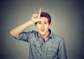 Young man showing loser sign on forehead, looking with disgust