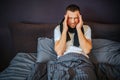 Young man sits on bed and holds hands close to forehead. He is suffering from headache. Pain is strong and terrible. Guy