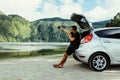 Young man sits in the car trunk take selfie photo on phone near beauty lake background. Summer vocation. Car trip Royalty Free Stock Photo