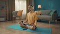 A young man sits cross legged in a lotus position with his eyes closed on a sports mat next to a smartphone. A man in a