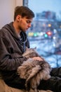 Young man sits at home by the window and holds fat fluffy cat in his sleeve Royalty Free Stock Photo