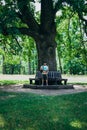 A young man is sitting on a bench near a tall tree. Man on a bench in a green park. Handsome young man in shorts on a wooden bench