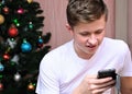 young man is sitting by Christmas tree, looking at phone and smiling. Watch videos and exchange text messages on your smartphone. Royalty Free Stock Photo